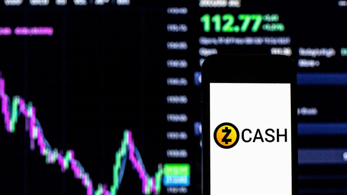 A phone with a Zcash logo is held up in front of a computer monitor displaying price information